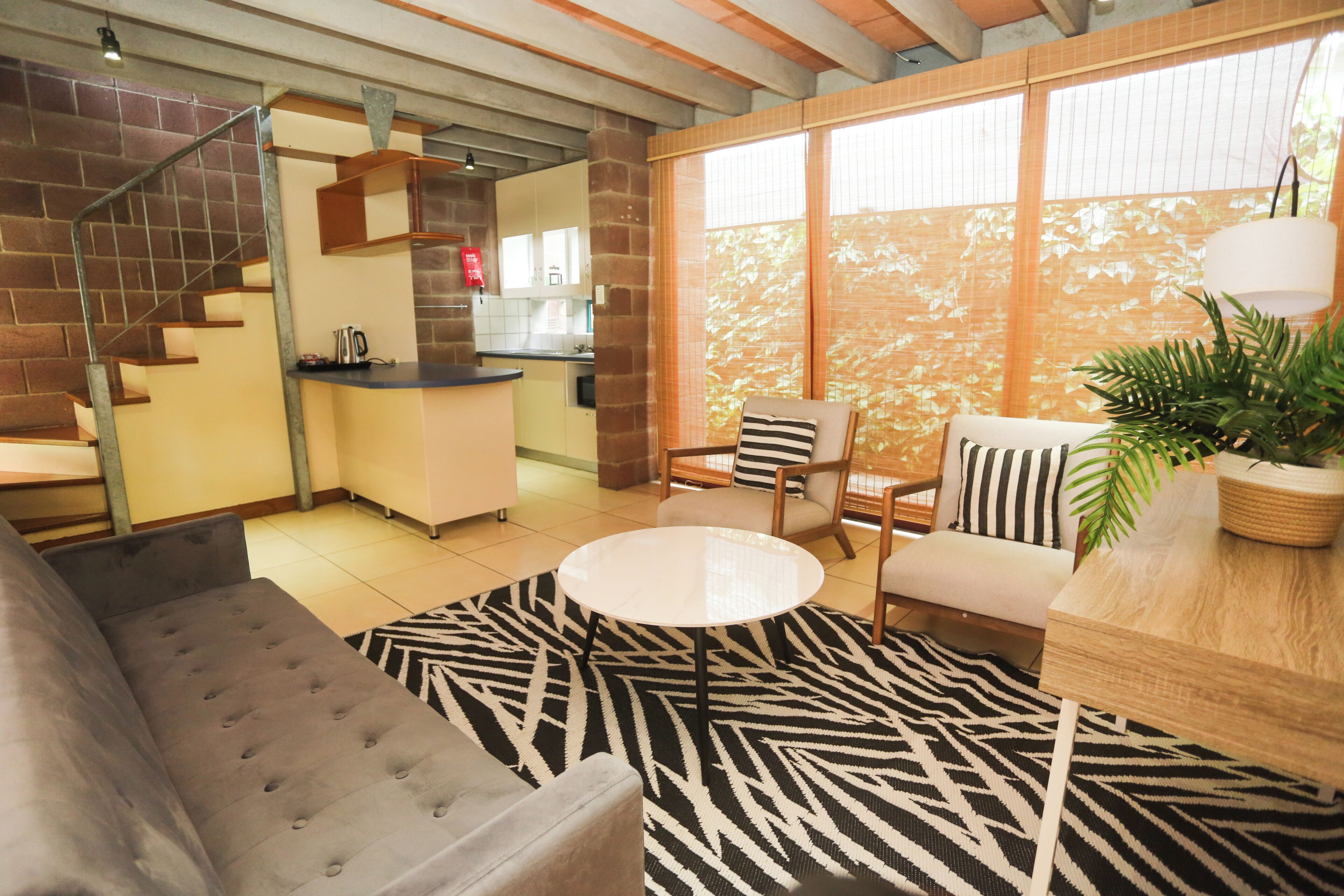 A modern living room with a sofa, a round coffee table, and a chair. There is a kitchen area with a counter and cabinets visible through an open door.