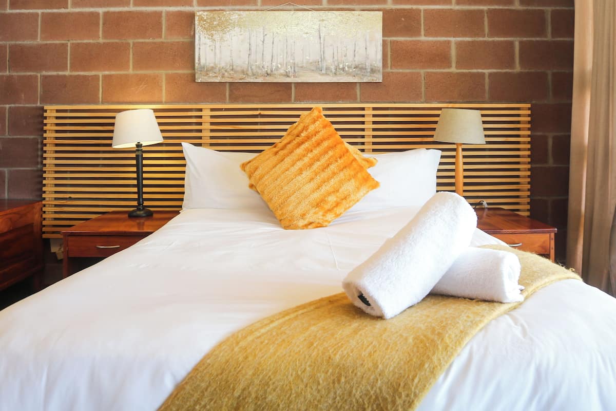 Una cama bien tendida con cabecera de madera, dos lámparas de noche y un cojín decorativo.