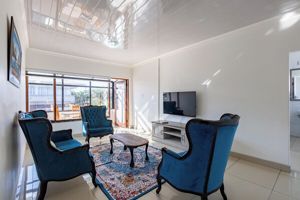 A living room with a TV, blue chairs, a coffee table, and a patterned rug. - Safi Self-Catering Suites (East London)