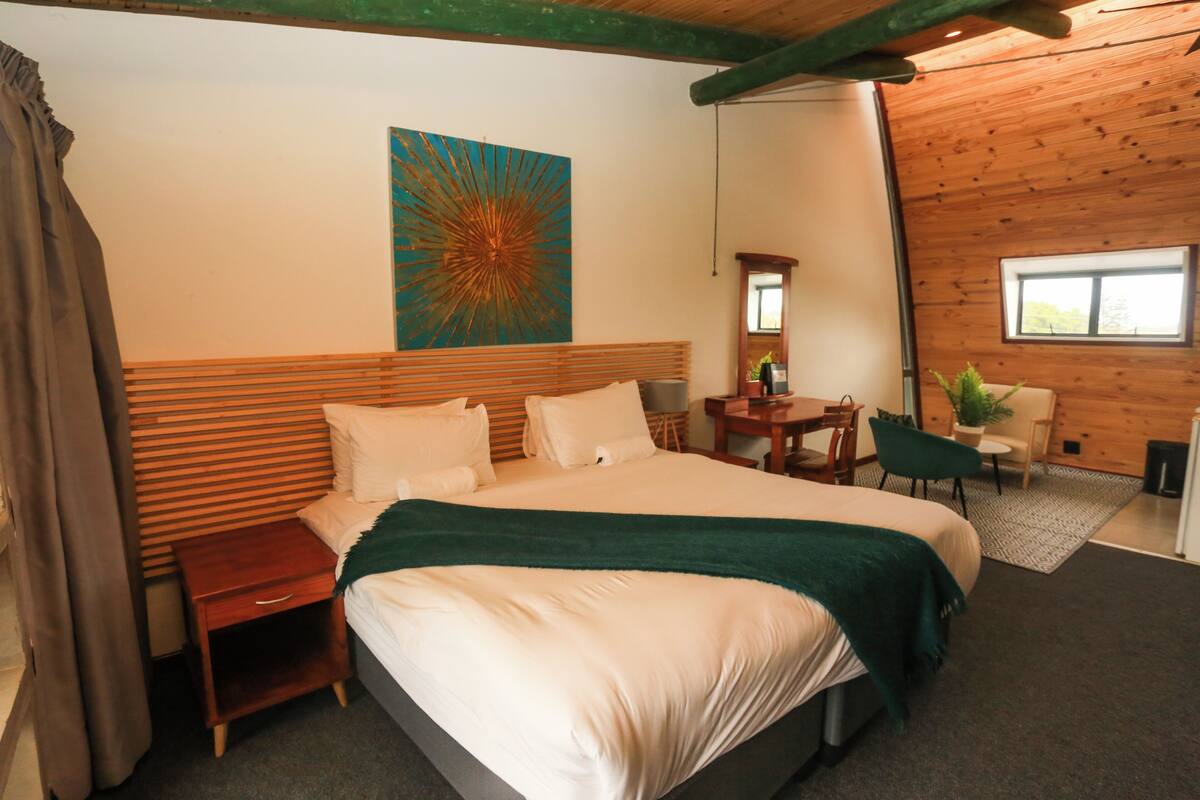 Una habitación de hotel con una cama grande, un cabecero de madera, un escritorio con una silla, una mesita con otra silla y una ventana.