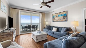 A spacious living room with a balcony overlooking the sea, a ceiling fan, a mounted TV, and a beach-themed wall art. - Ocean Ritz 2 Primary Suites | Expansive Balcony | Sleeps 12 (Panama City Beach)