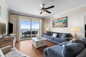 Ein geräumiges Wohnzimmer mit Balkon und Meerblick, Deckenventilator, Wand-TV und Strandbilder an den Wänden.