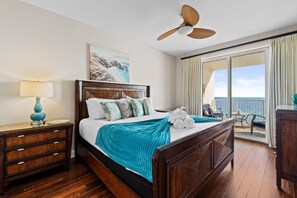 Ein Hotelzimmer mit Bett, Nachttisch, Deckenventilator, einem Gemälde und einem Balkon mit Meerblick.