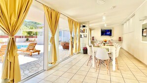 A modern living room with a dining area, large windows with curtains, a pool view, and a flat-screen TV on the wall. - Sidari Beach Villa with private pool (Sidari)
