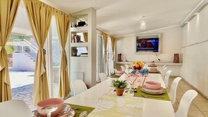 A dining area with a long table set for four, a television mounted on the wall, and a staircase leading to an outdoor area. - Sidari Beach Villa with private pool (Sidari)