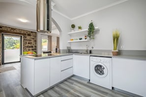 A modern kitchen with white cabinetry, a built-in washing machine, a stone accent wall, and a window with a view. - Basalt House A by Madeira Sun Travel (São Vicente)