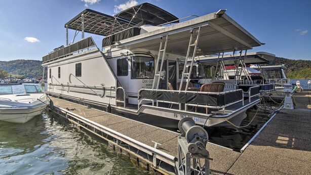 A houseboat docked at a marina with a covered deck area, a small cabin, and a motorized outboard.