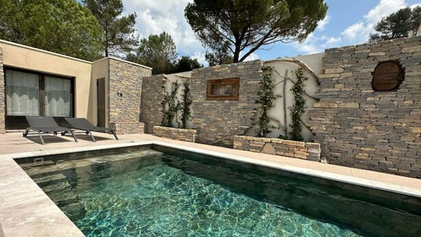 A swimming pool with clear water, surrounded by stone walls and a modern building with a covered patio. - Villa Avec Piscine Loft Cabane (Nîmes)