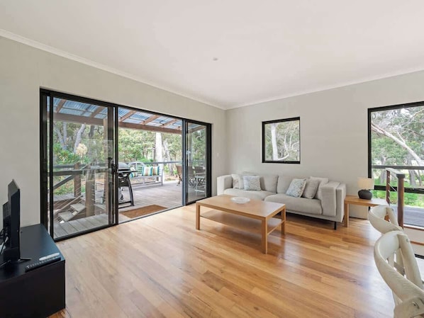 A spacious living room with a wooden floor, a sofa, a coffee table, and large sliding glass doors leading to an outdoor patio.