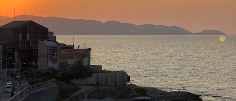 A coastal sunset view with a promenade, buildings, and a calm sea.