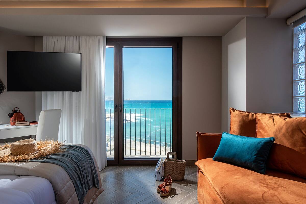 A modern hotel room with a large balcony, a bed with a view, a flat-screen TV, and a cozy seating area.