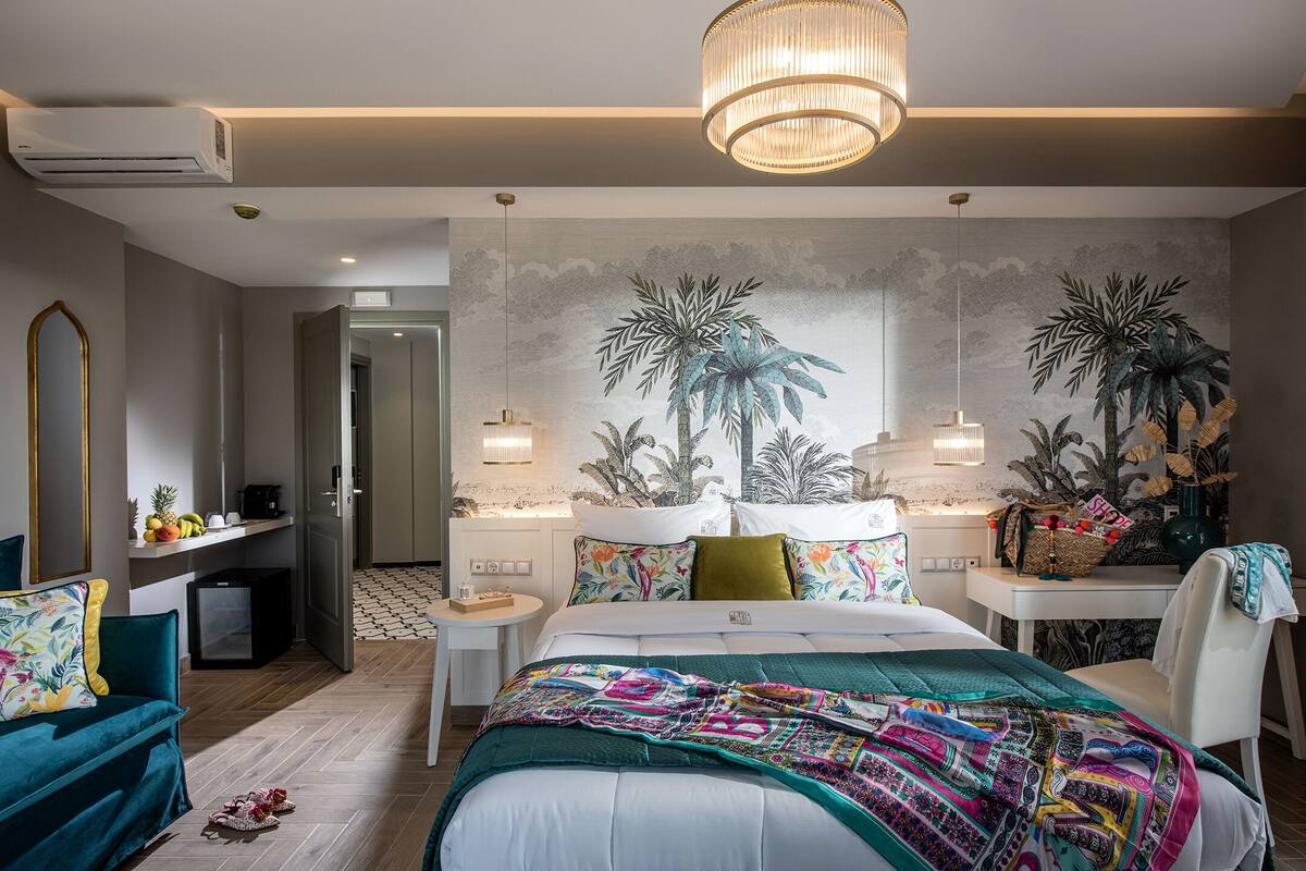 A modern hotel room with a large bed, a desk, a chair, and a small table. There is a wall mural of tropical plants, a chandelier, and an air conditioning unit.