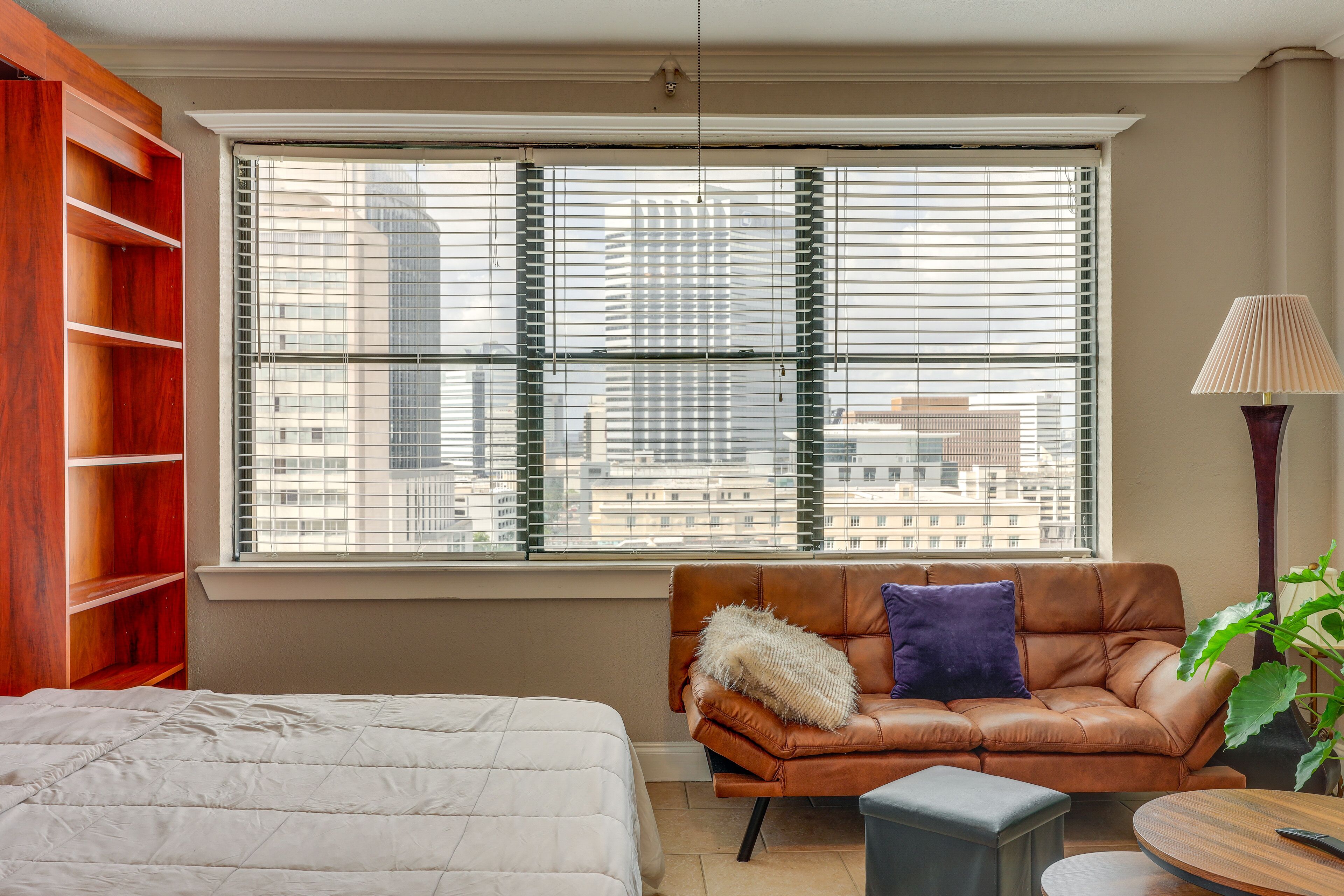 A bedroom with a large window, a brown leather sofa, a bedside table, and a bed with a white duvet.