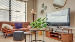 A living room with a brown leather sofa, a round coffee table, a TV on a wooden stand, a potted plant, and a lamp. - Downtown Jacksonville Studio w/ City Views! (Jacksonville)