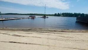 Une plage de sable fin bordée d’un lac paisible, un quai avec des bateaux, et une côte lointaine parsemée d’arbres.