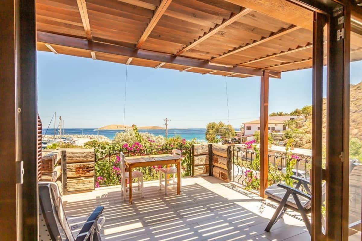 A covered outdoor area with a wooden table and chairs, a bench, and a view of the sea and hills.