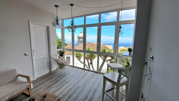 Un balcon avec vue sur la mer et les bâtiments, une porte blanche, un fauteuil suspendu et une petite table.