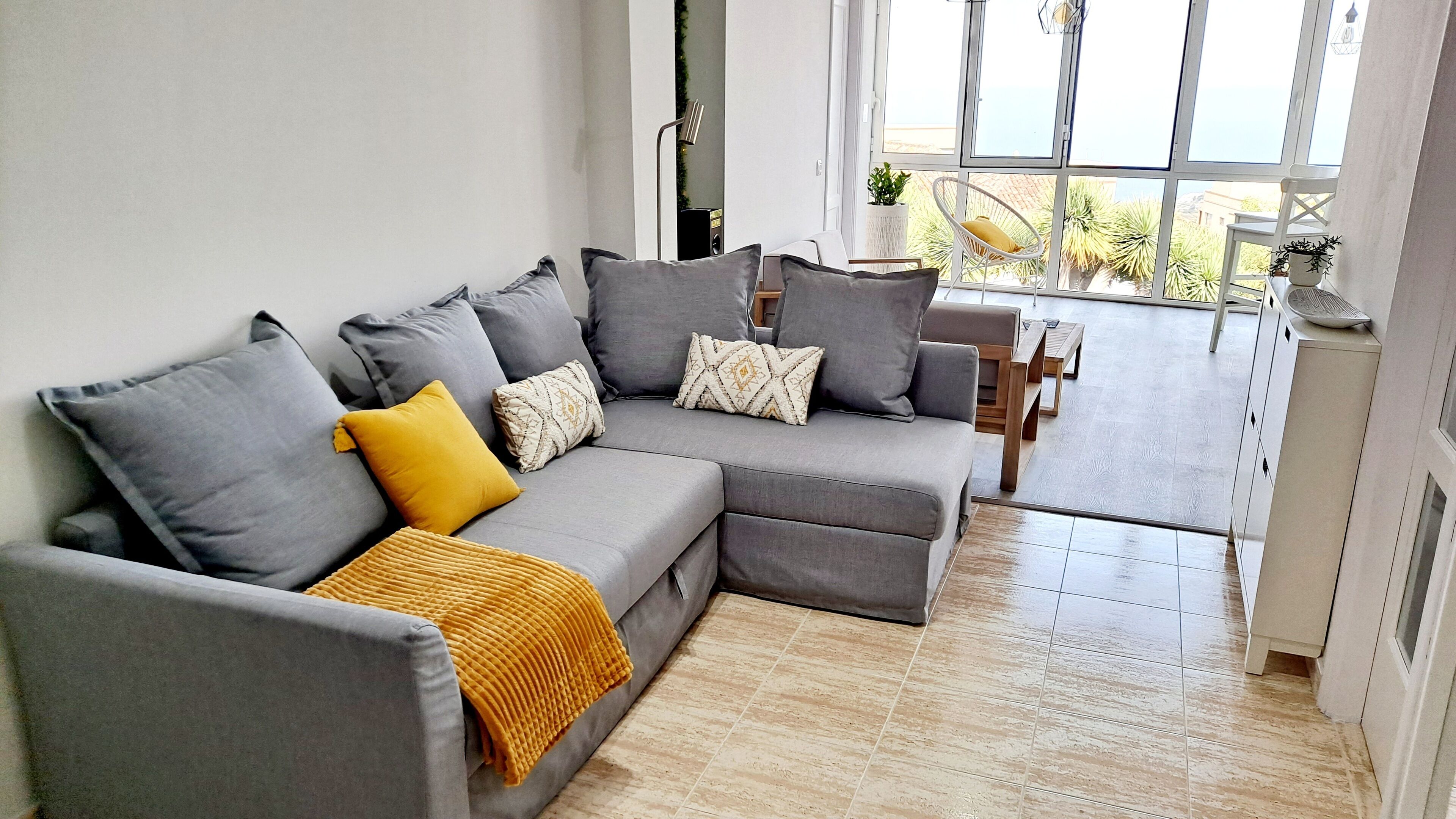 A modern living room with a grey sectional sofa, a yellow throw blanket, and a view of the outdoors through large windows.