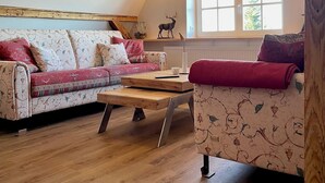Une pièce confortable de style loft, avec des poutres apparentes, un canapé, un fauteuil et une fenêtre donnant sur un paysage verdoyant.