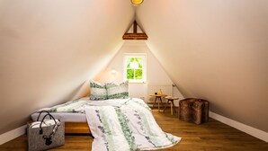 Une chambre mansardée chaleureuse, avec un plafond en pente, un lit recouvert d’une literie à motifs floraux, une petite table en bois et une fenêtre donnant sur un paysage verdoyant.