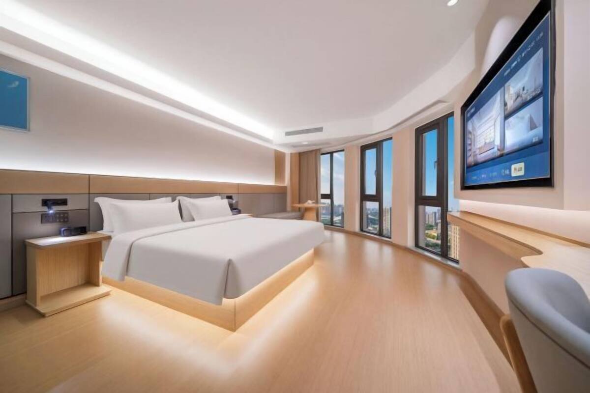 A modern hotel room with a large bed, a TV mounted on the wall, and a view of the city through large windows.