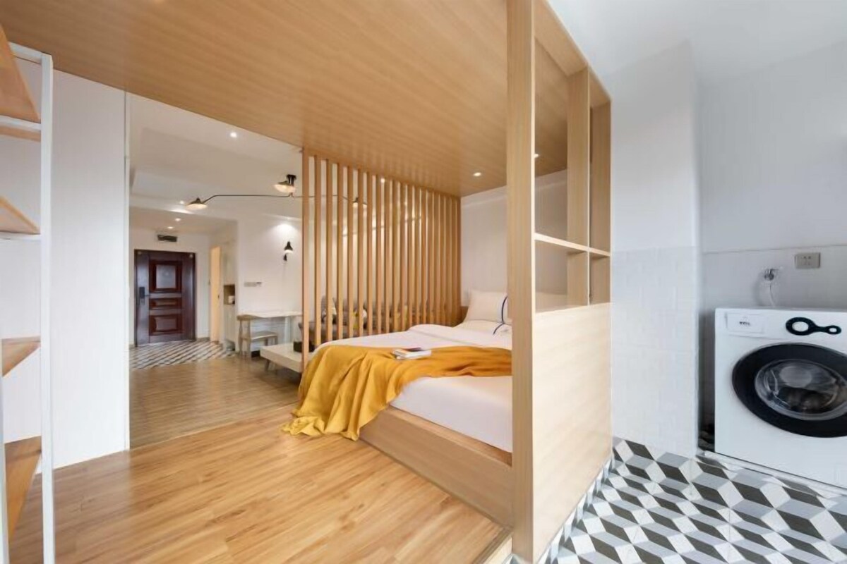 A modern hotel room with a wooden floor, a bed with a yellow blanket, a washing machine, and a patterned floor.
