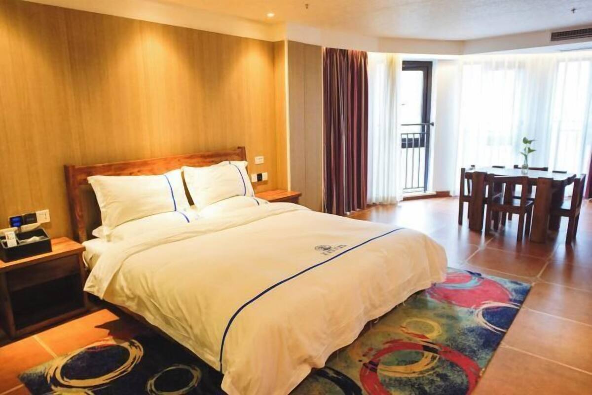 A hotel room with a large bed, a wooden headboard, a desk with chairs, and a balcony with curtains.