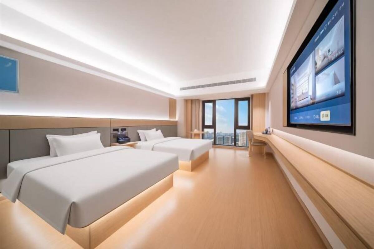 A modern hotel room with two beds, a large TV screen, and a view of the city.
