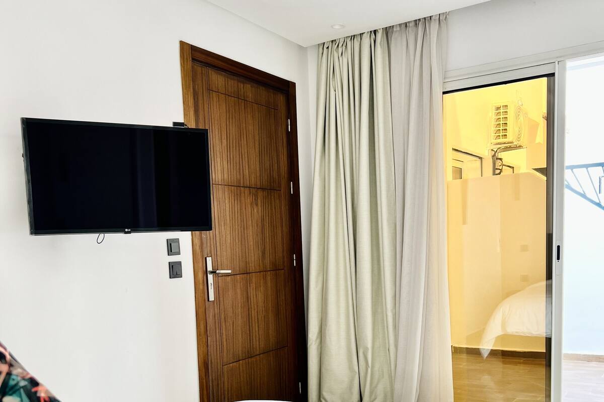 Habitación de hotel con cama, televisor montado en la pared, puerta de madera y puerta corrediza de vidrio que da a un balcón.