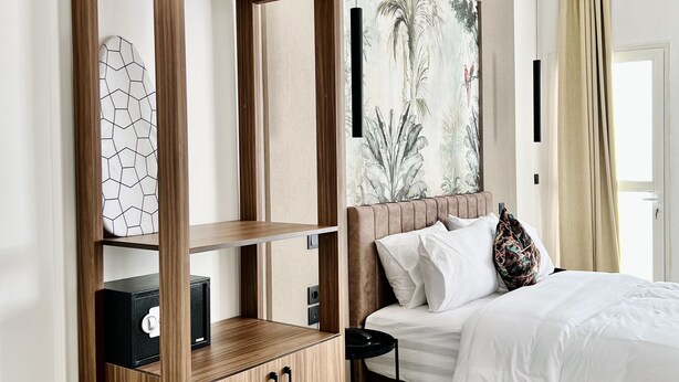 A modern hotel room with a wooden cabinet, a bed with white bedding, a large abstract painting, and a view of palm trees through the window. - MyCity (Rabat)
