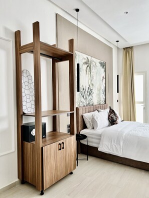 Une chambre d’hôtel moderne avec un meuble en bois, un lit recouvert de linge de maison blanc, un grand tableau abstrait et une vue sur les palmiers par la fenêtre.