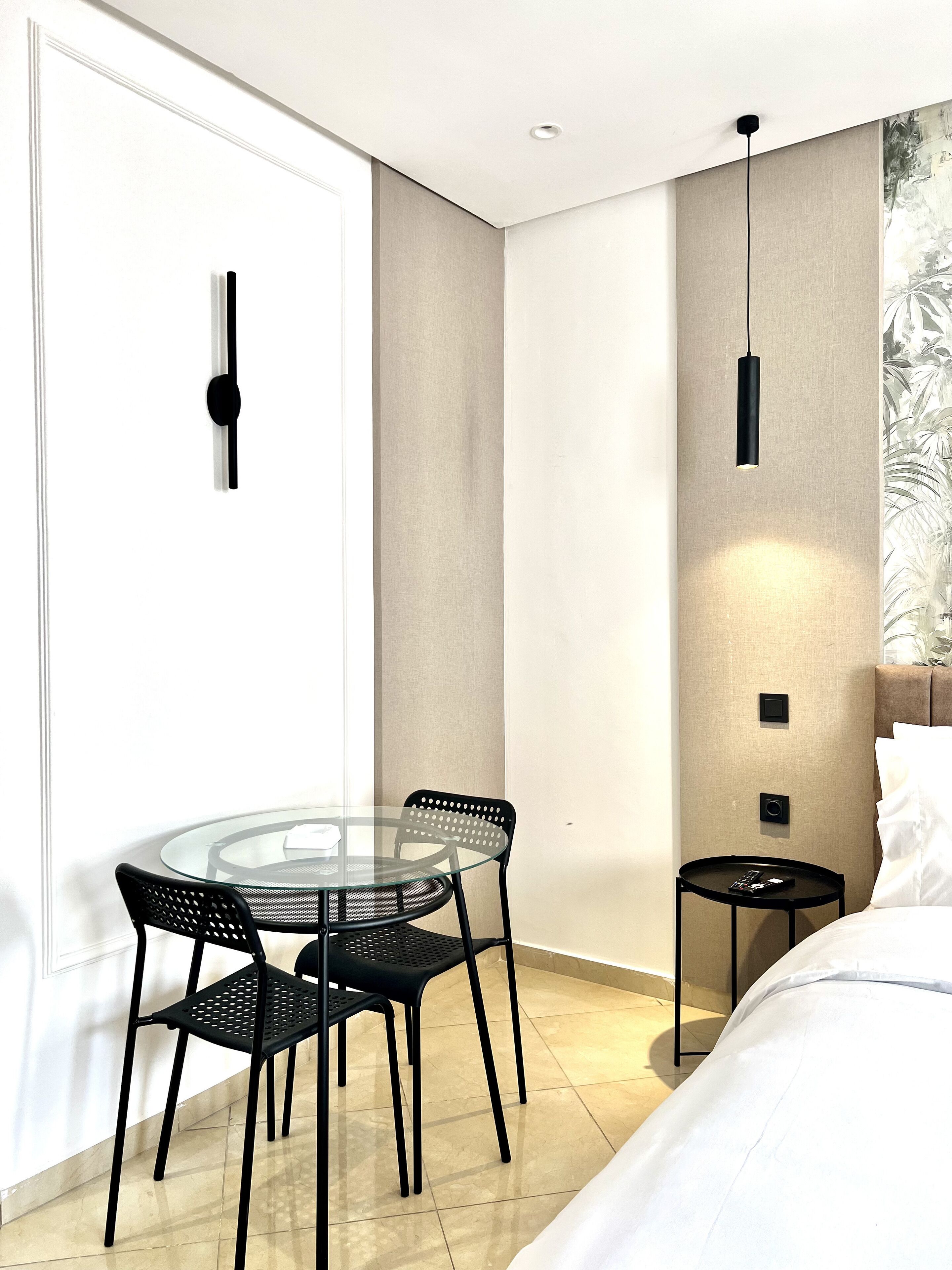 Una habitación de hotel moderna con una mesa y sillas de vidrio, una cama, una mesita de noche y una lámpara fijada a la pared.
