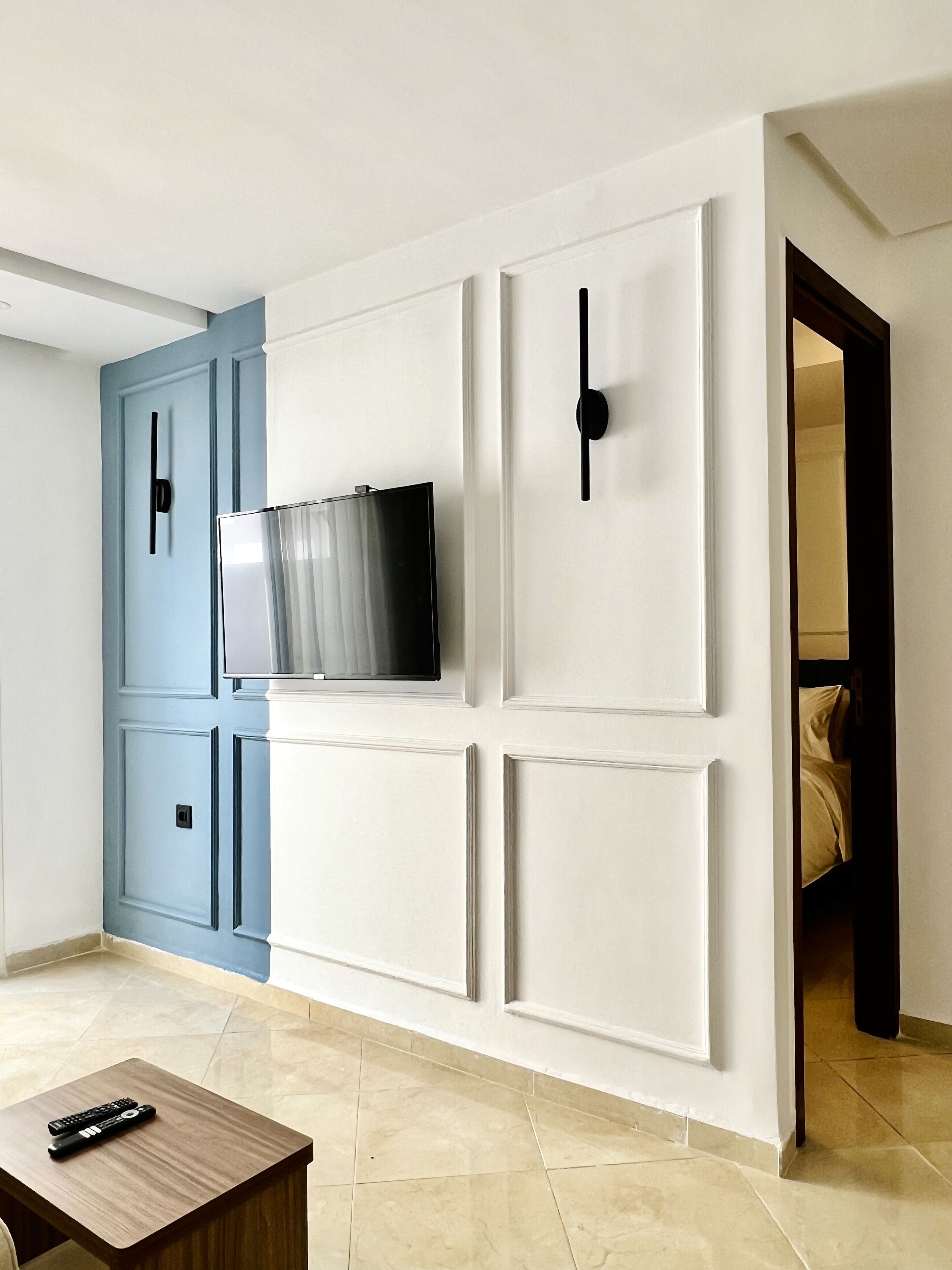 A modern living room with a flat-screen TV mounted on a white wall, a blue door, and a wooden coffee table with remote controls.