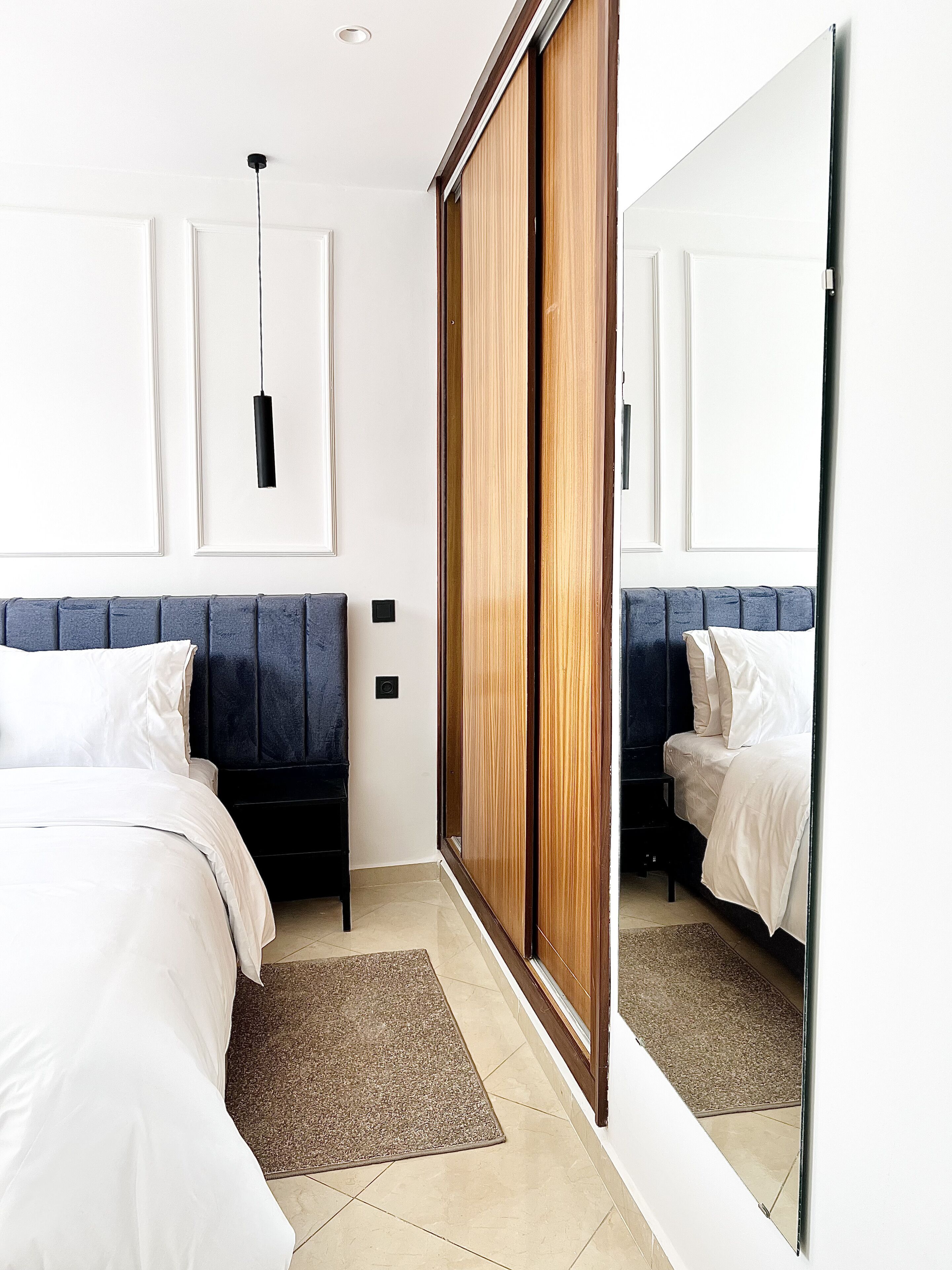 A modern bedroom with a large mirror, a bed with white linens, a dark blue headboard, and a wooden wardrobe.
