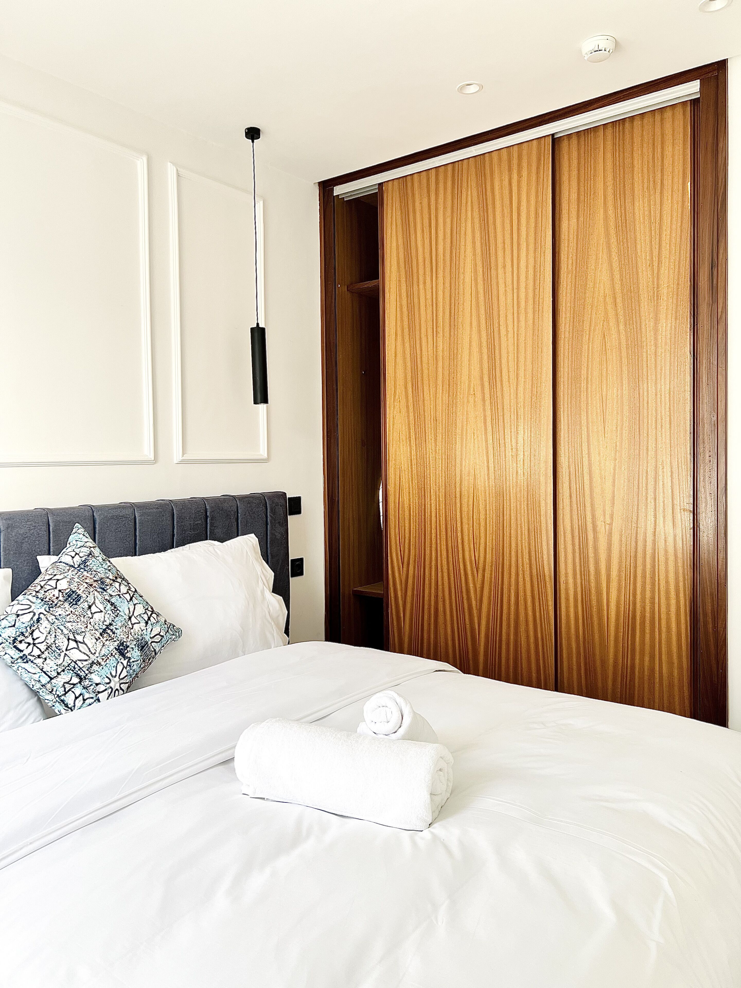 Un lit bien fait, avec du linge de lit blanc et une serviette enroulée, une tête de lit rembourrée de couleur grise et une armoire en bois dotée d’une porte coulissante.