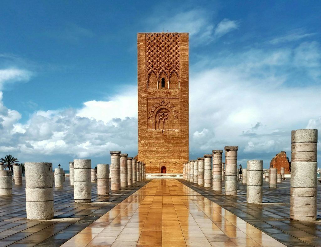 Een hoge bakstenen toren met een gedetailleerd gevelontwerp, omringd door gelijkmatig geplaatste kolommen op een verharde looppad.