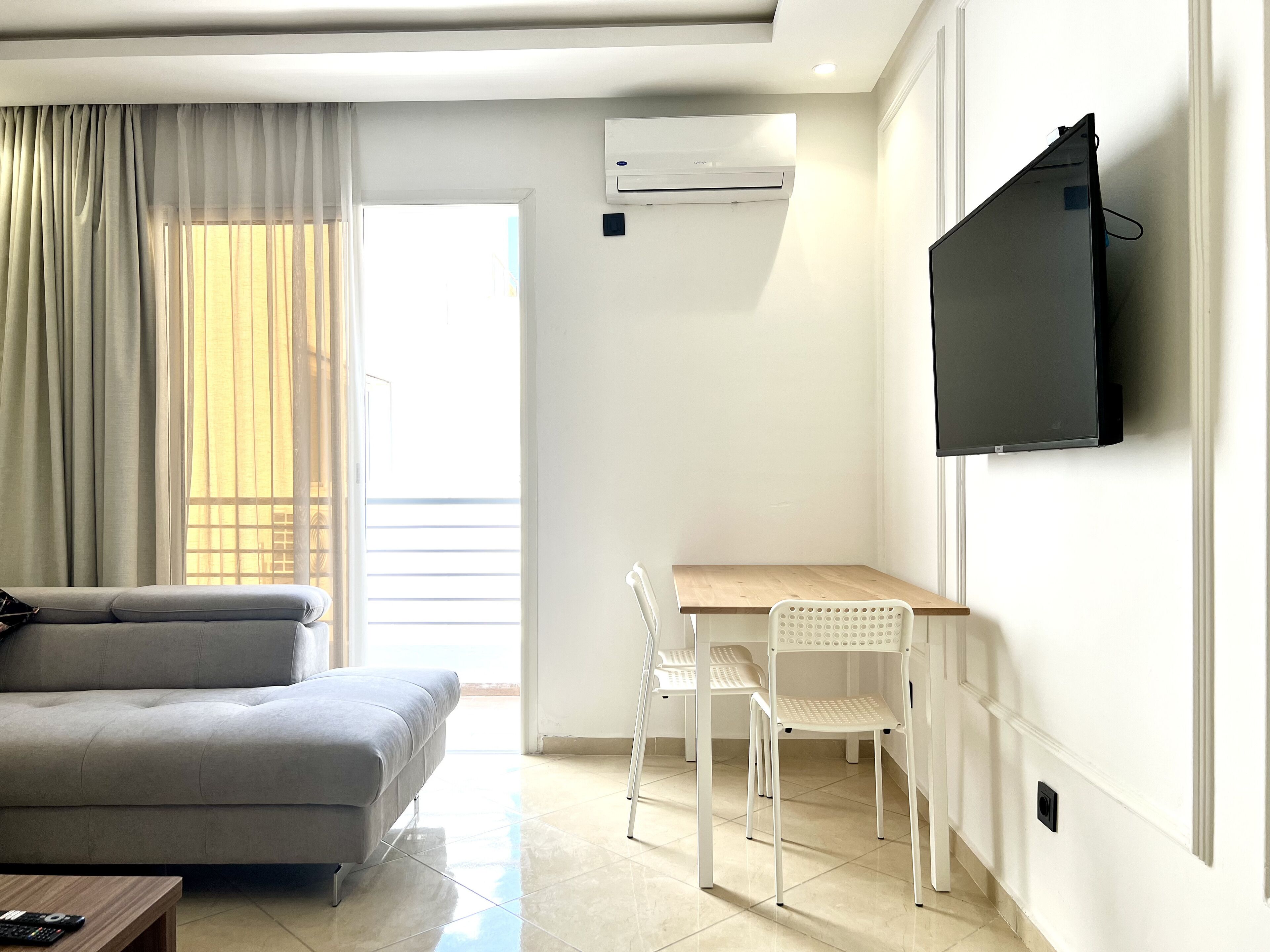 Uma sala de estar moderna com um sofá cinzento, uma mesa de madeira e uma cadeira branca. Há uma televisão de tela plana na parede e um ar-condicionado acima.