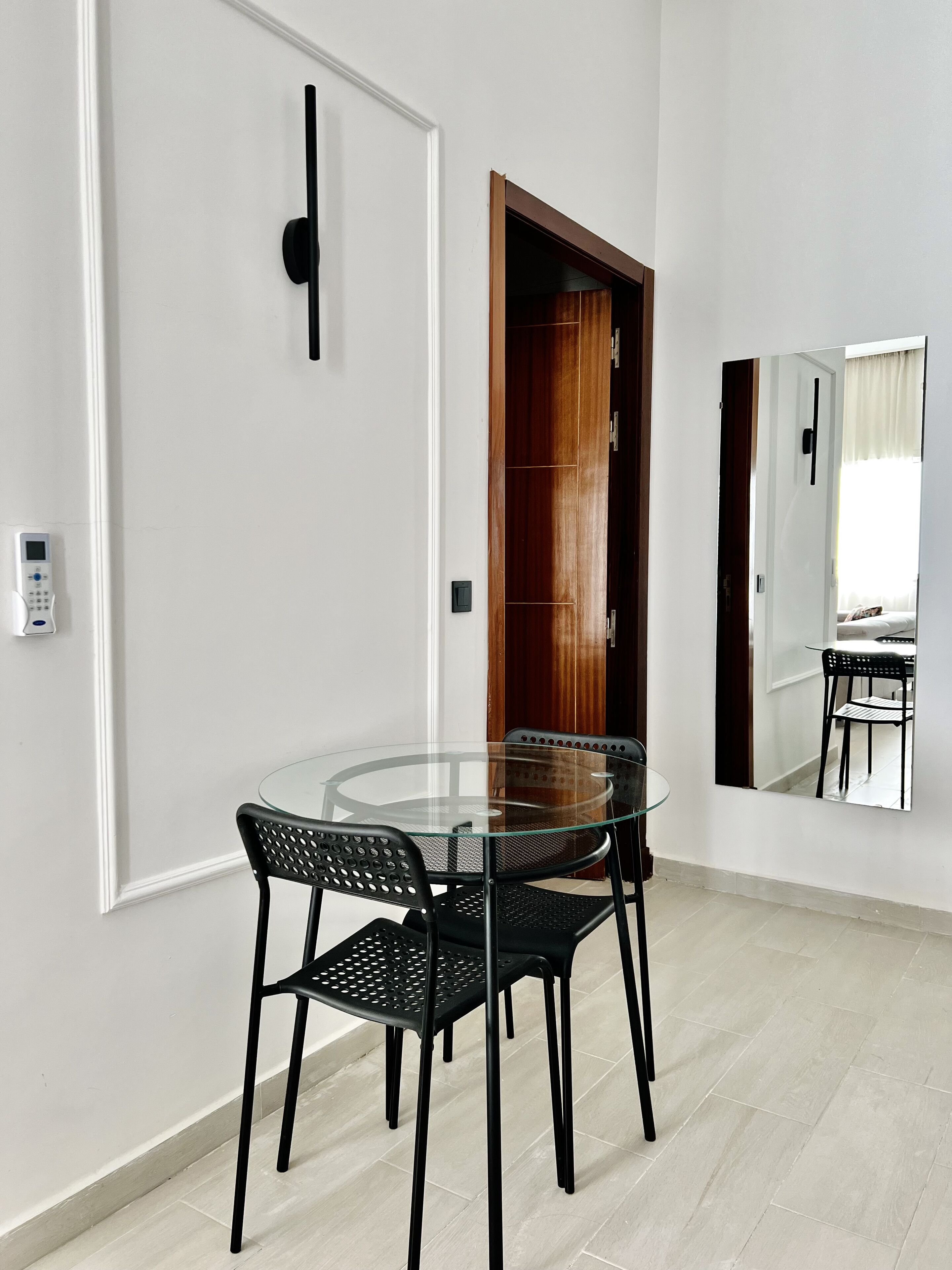 A modern dining area with a glass table and black chairs, a wooden door, and a white wall with a doorbell.