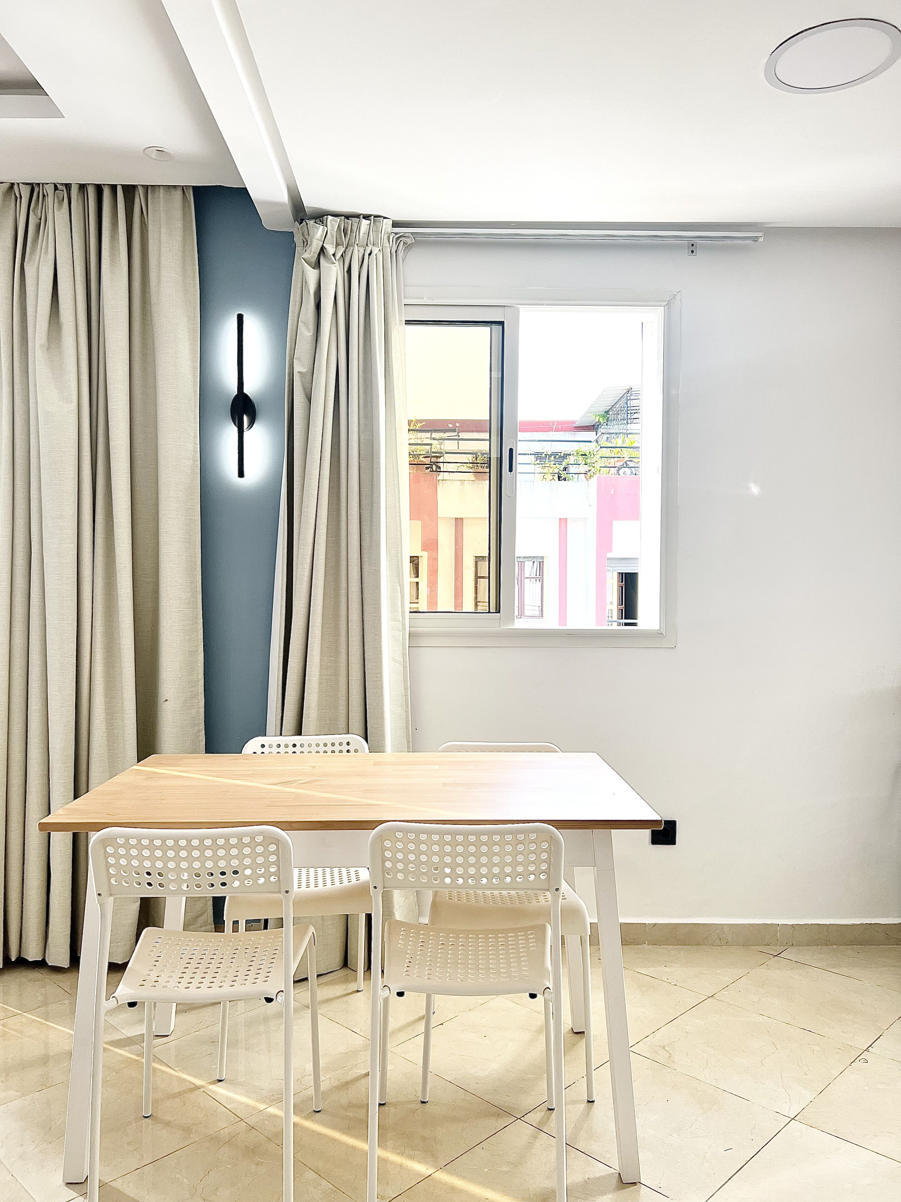 Une pièce avec une table en bois et des chaises blanches, une grande fenêtre donnant sur des immeubles et un rideau sur le côté gauche.
