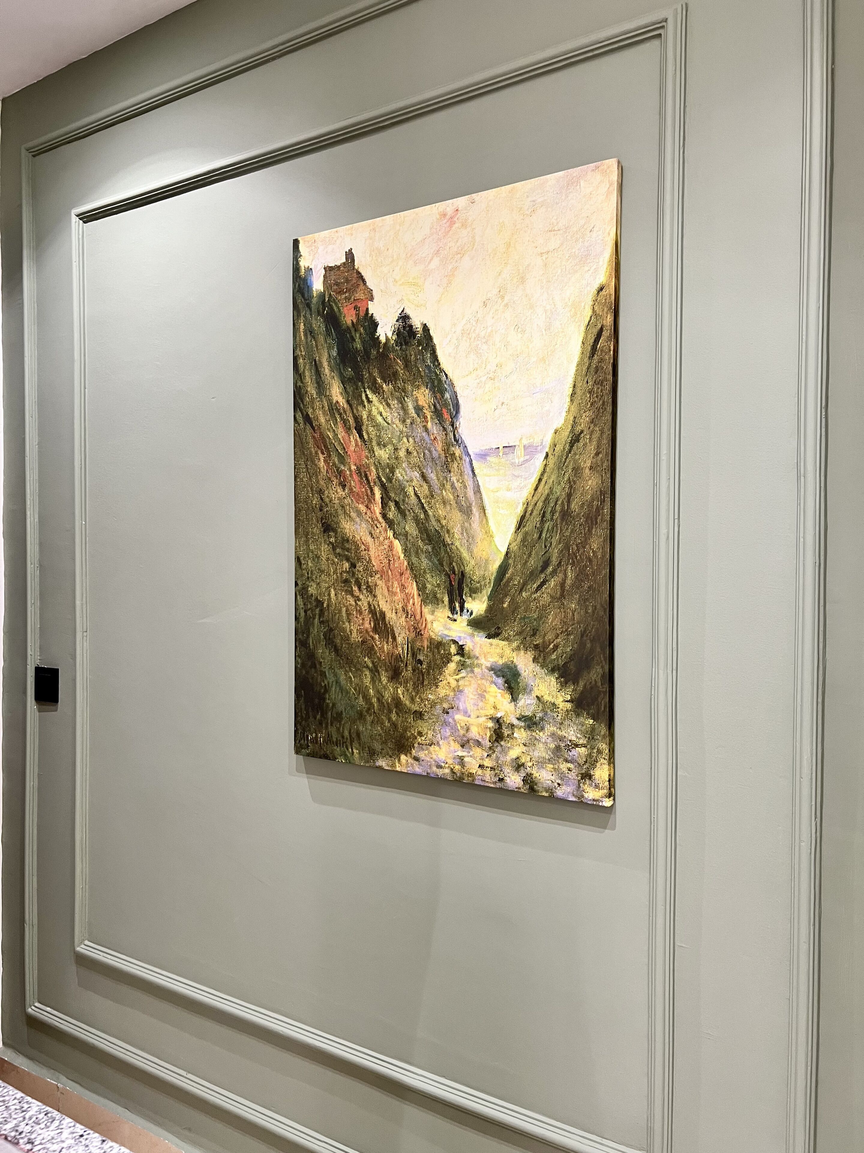 A painting of a canyon with a path and a bridge in the distance, framed and hung on a wall.