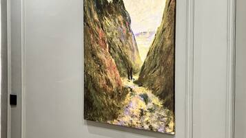 A painting of a canyon with a path and a bridge in the distance, framed and hung on a wall.