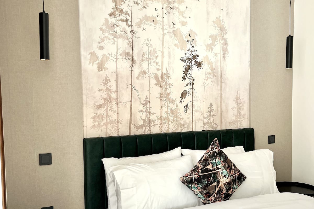 Un dormitorio con una cama, un cabecero tapizado en verde, ropa de cama blanca, una almohada decorativa y una mesita de noche negra, además de una pared con un mural de un bosque.