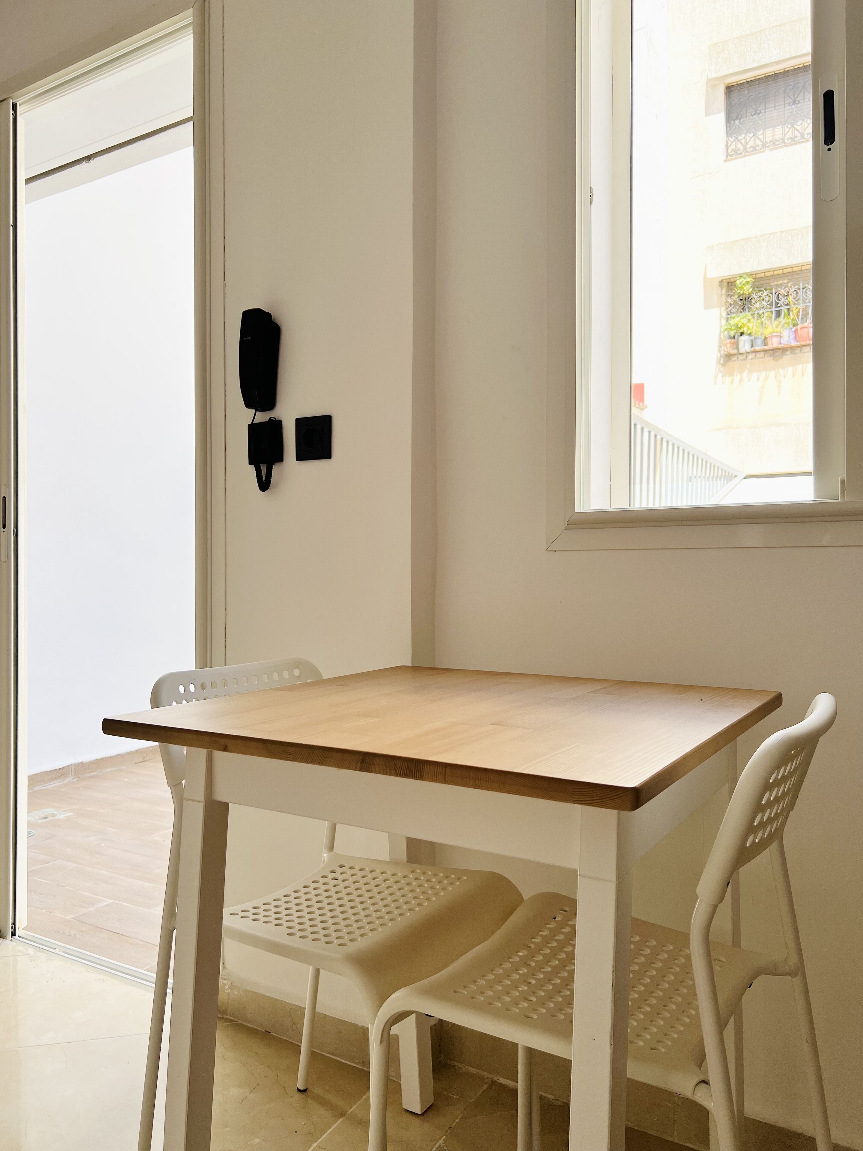 A room with a wooden table and two white chairs, a phone on the wall, and a window with a view of the outside.