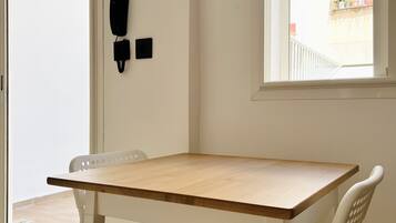 A room with a wooden table and two white chairs, a phone on the wall, and a window with a view of the outside.