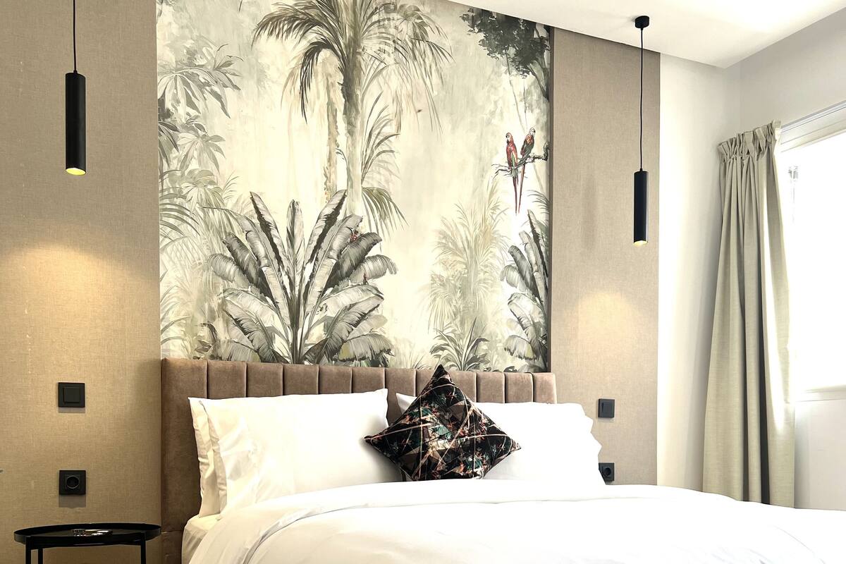 Un dormitorio moderno con una cama grande, cabecera con un mural tropical, una mesita de noche pequeña y un ventanal con cortinas.