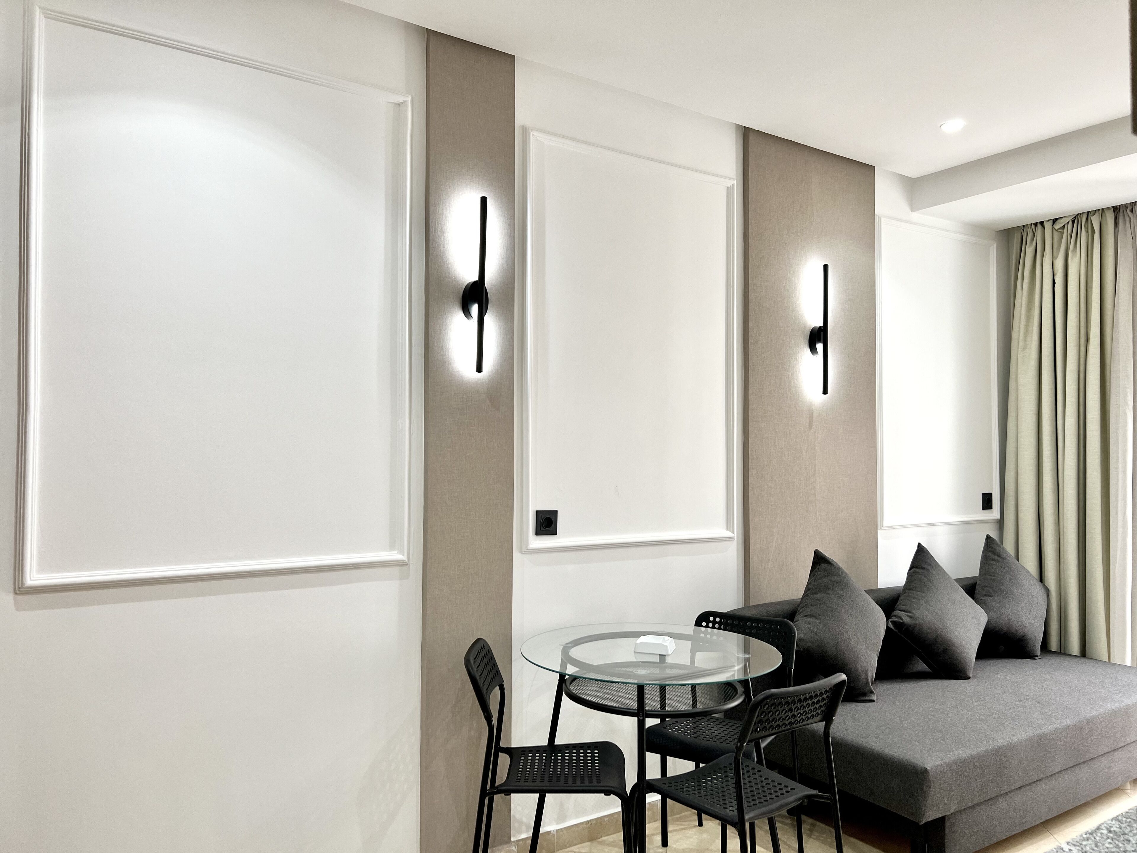 Una sala de estar moderna con una mesa de vidrio, sillas negras, un sofá gris con cojines e iluminación montada en la pared.