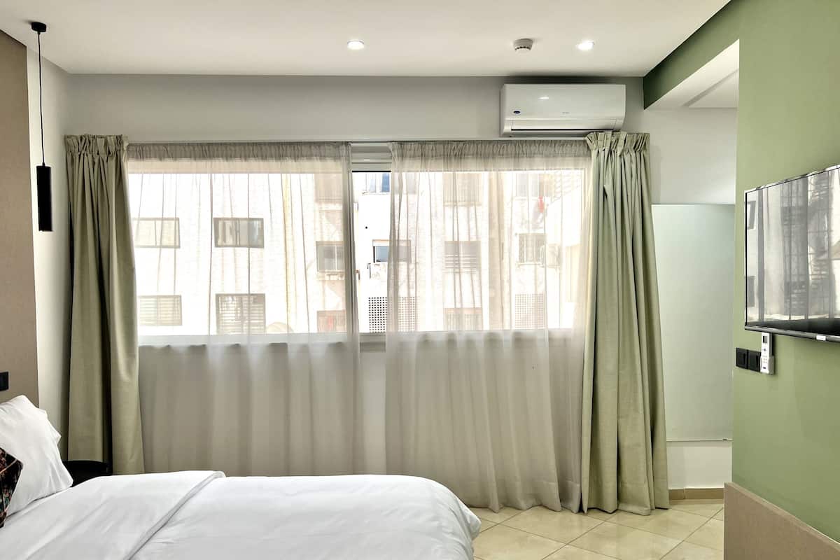 Habitación de hotel con cama, cortinas, aire acondicionado, televisor y vistas a edificios desde la ventana.
