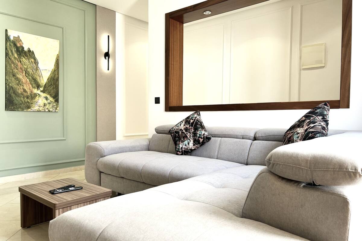 Una sala de estar moderna con un sofá seccional gris, dos cojines estampados, una mesa de centro de madera con un control remoto y un cuadro de paisaje en la pared.