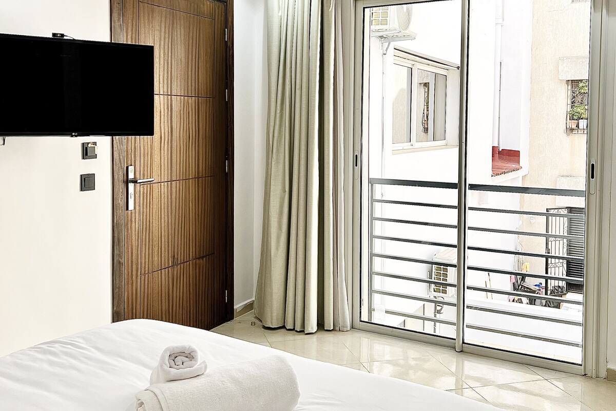 Habitación de hotel con una cama, un televisor montado en la pared, una puerta de madera y un ventanal corredero que da a un balcón.
