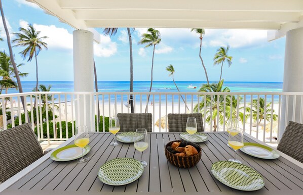 Ein Tisch für vier Personen, gedeckt mit Tellern, Gläsern und einem Korb mit Croissants, mit Blick auf einen Strand mit Palmen und dem Meer.